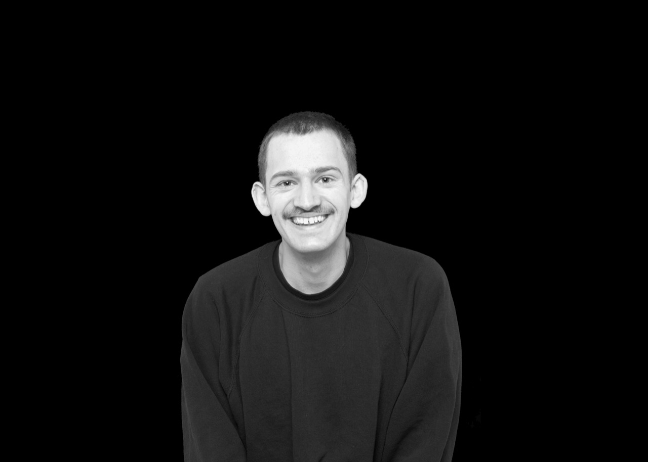 Porträt von Jonas Nitzl: lächelnd, im dunklen Pullover vor schwarzem Hintergrund